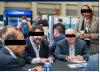 Photobombed with diversity Three men (two very white) in similar color suit jackets seated at a table with a woman of color in the edge of the image, standing, in a different hued jacket.